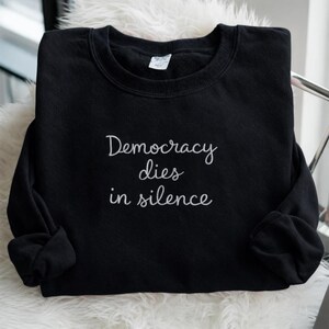 May include: Black sweatshirt with the embroidered phrase "Democracy dies in silence" in white script. The long-sleeved crewneck is folded neatly, showcasing the message prominently. The sweatshirt is displayed on a white, fluffy surface.