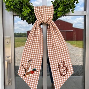 May include: A brown and white gingham fabric door hanger featuring a green wreath at the top. One side displays an embroidered mallard duck in flight, while the other side features a brown embroidered script monogram of the letter B.