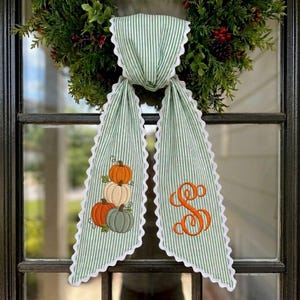 May include: A green and white striped fabric door hanger featuring a scalloped white edge. One side displays an embroidered stack of three pumpkins in orange, white, and sage green. The other side features an embroidered orange letter S.