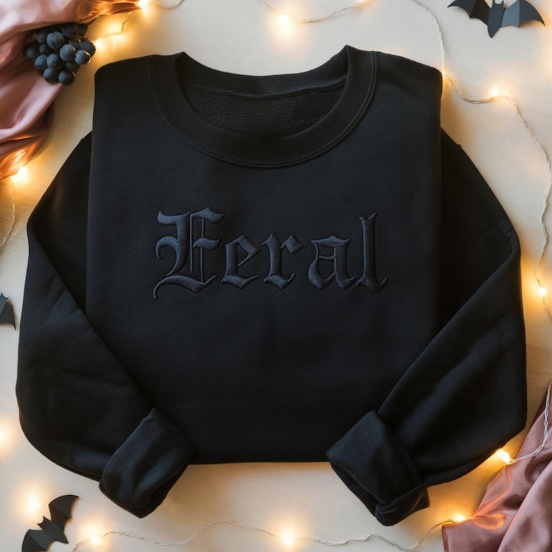 May include: Black crewneck sweatshirt with the word "Feral" embroidered in gothic lettering across the chest. The long-sleeved top is made of a soft material. The sweatshirt is displayed on a light-colored surface with decorative lights and Halloween-themed cutouts.