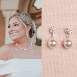 May include: A pair of silver earrings with a pearl drop and a crystal cluster. The earrings are on a pink fabric background.