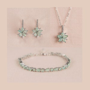 Könnte beinhalten: Ein silbernes Halsketten-, Armband- und Ohrring-Set mit einem floralen Design und hellgrünen Edelsteinen.
