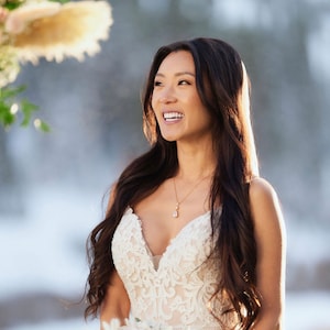 May include: A bride wearing a white lace wedding dress with a delicate gold necklace. The necklace is 18 inches long. She is holding a bouquet of blush pink and white roses.