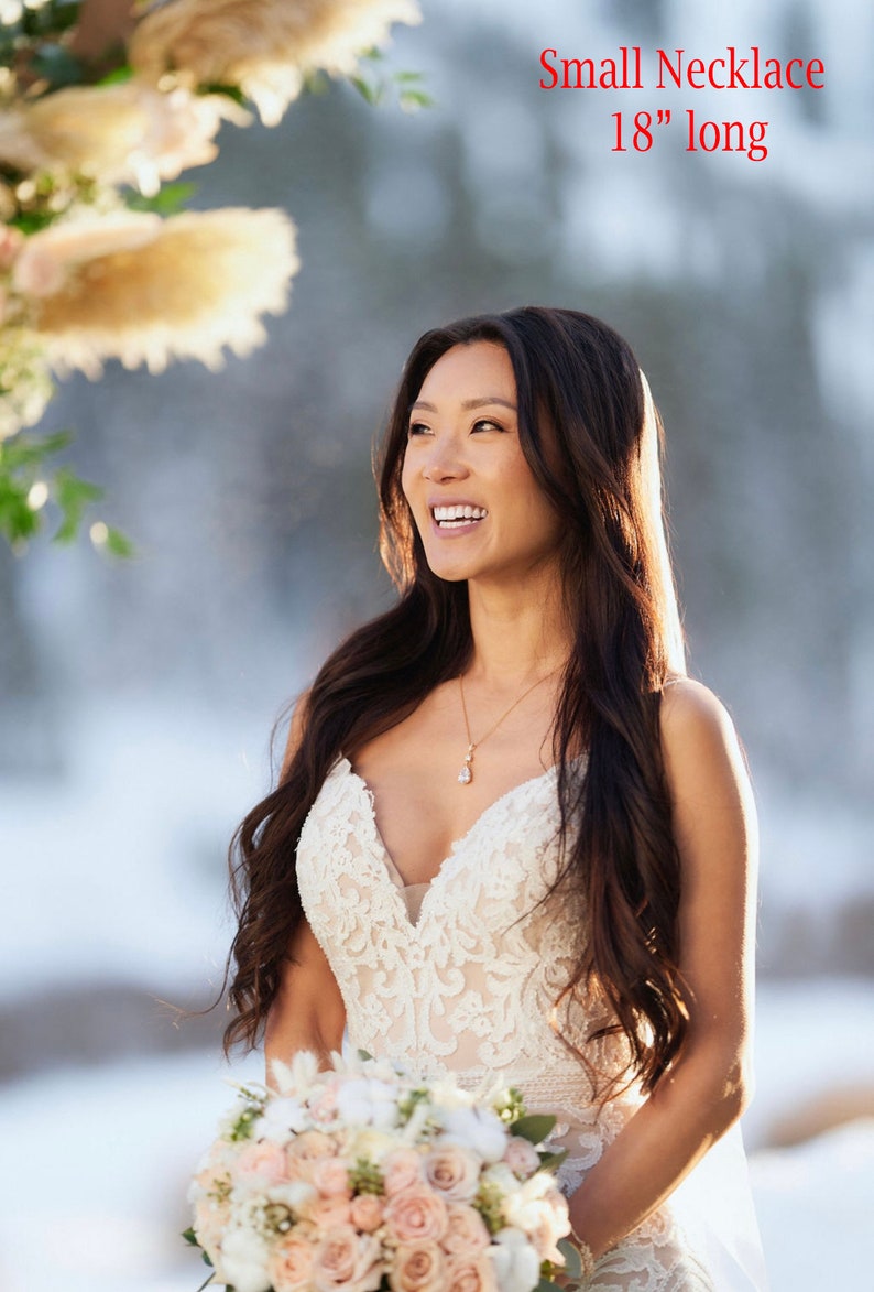 May include: A bride wearing a white lace wedding dress with a delicate gold necklace. The necklace is 18 inches long. She is holding a bouquet of blush pink and white roses.