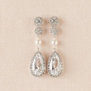 May include: Elegant silver-toned drop earrings. Each earring features a cluster of small, sparkling stones at the top, followed by a pearl, and a large, teardrop-shaped clear stone surrounded by smaller stones. The earrings are designed for formal occasions.