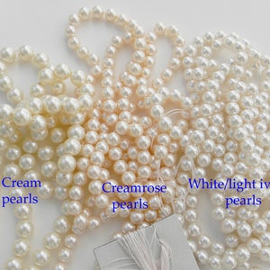 May include: Three strands of faux pearls in different shades of white: cream, creamrose, and white/light ivory. The pearls are arranged in a pile on a white surface.