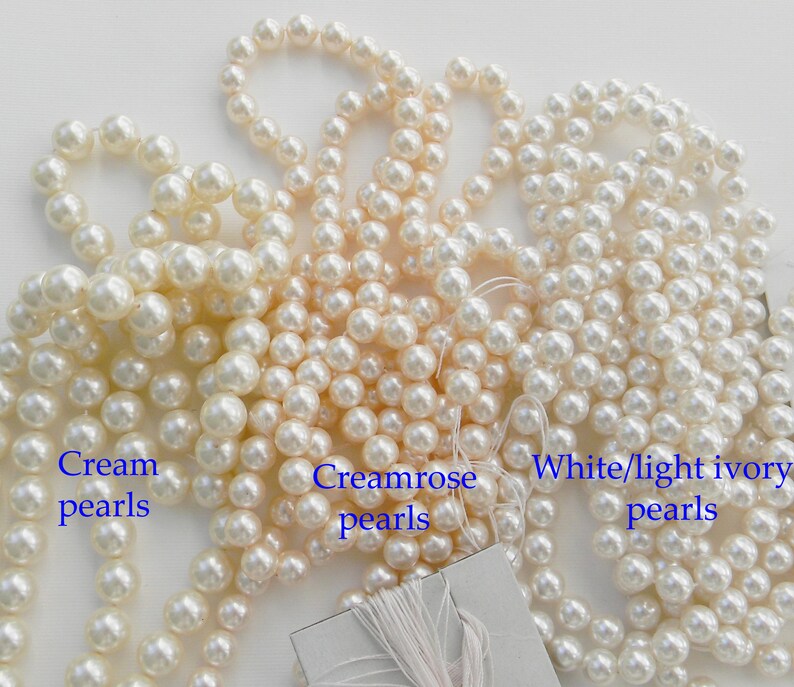 May include: Three strands of faux pearls in different shades of white: cream, creamrose, and white/light ivory. The pearls are arranged in a pile on a white surface.