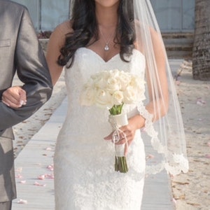 May include: A bride in a white lace wedding dress with a veil and a bouquet of white roses.