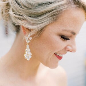 May include: A woman wearing large, gold, teardrop-shaped earrings with clear crystal accents. The earrings are dangling and have a floral design at the top.