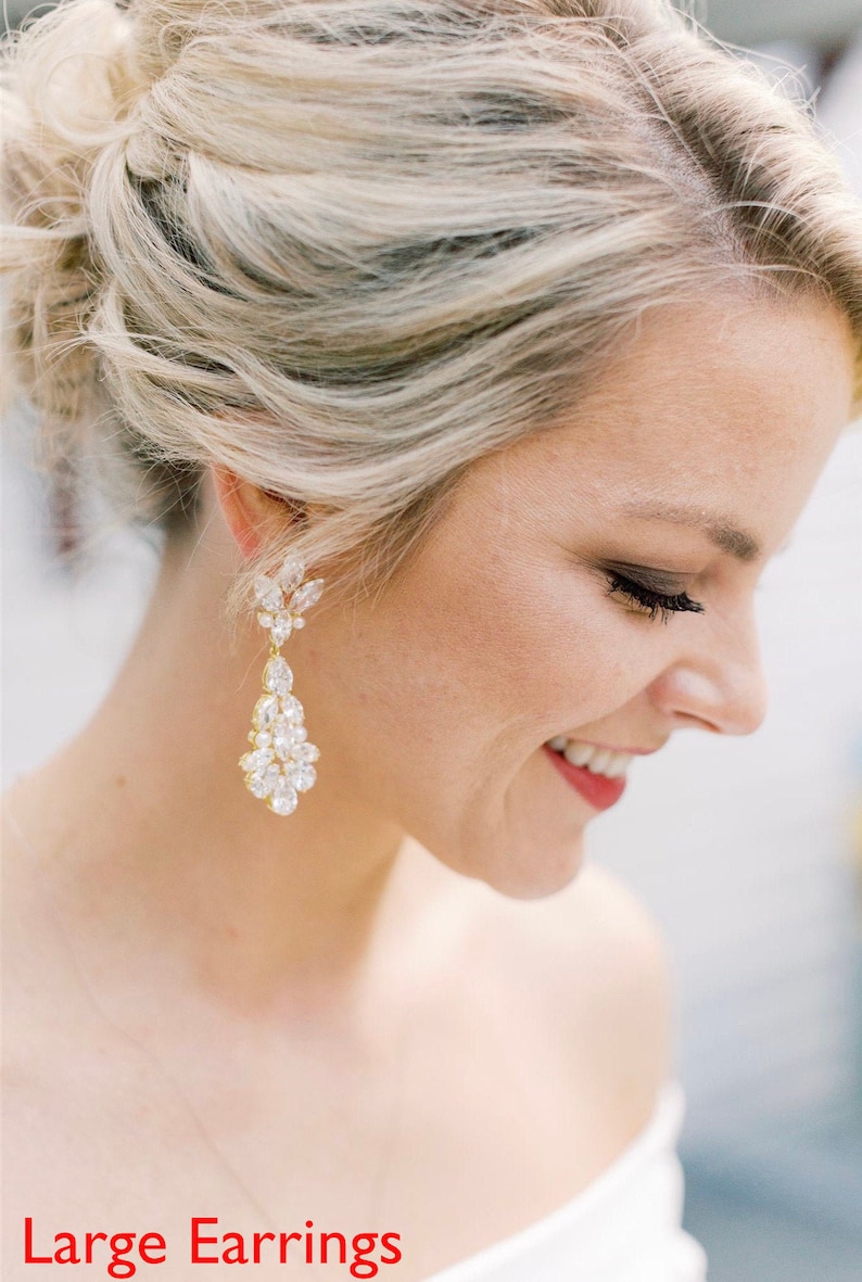 May include: A woman wearing large, gold, teardrop-shaped earrings with clear crystal accents. The earrings are dangling and have a floral design at the top.