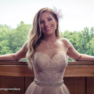 May include: A woman wearing a strapless, lace wedding dress with a beaded belt. She is standing in front of a window with a floral hairpiece and a necklace.