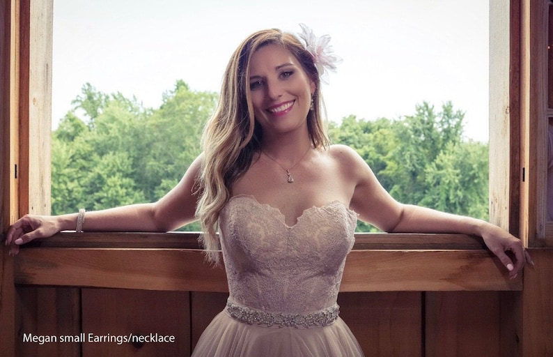May include: A woman wearing a strapless, lace wedding dress with a beaded belt. She is standing in front of a window with a floral hairpiece and a necklace.