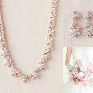 May include: A rose gold necklace and earrings set with clear cubic zirconia stones arranged in a floral pattern. The necklace is a delicate chain with a floral design and the earrings are dangle earrings with a floral design.