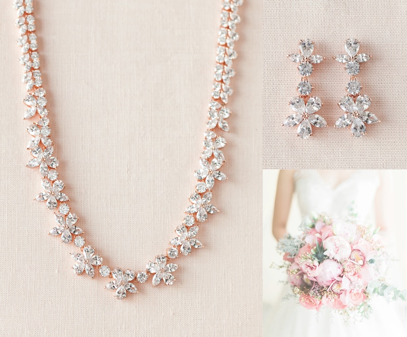 May include: A rose gold necklace and earrings set with clear cubic zirconia stones arranged in a floral pattern. The necklace is a delicate chain with a floral design and the earrings are dangle earrings with a floral design.