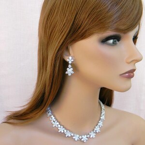 May include: A silver necklace and earrings set with clear crystal flower accents. The necklace is a delicate chain with a row of flower-shaped crystals. The earrings are drop earrings with three flower-shaped crystals.