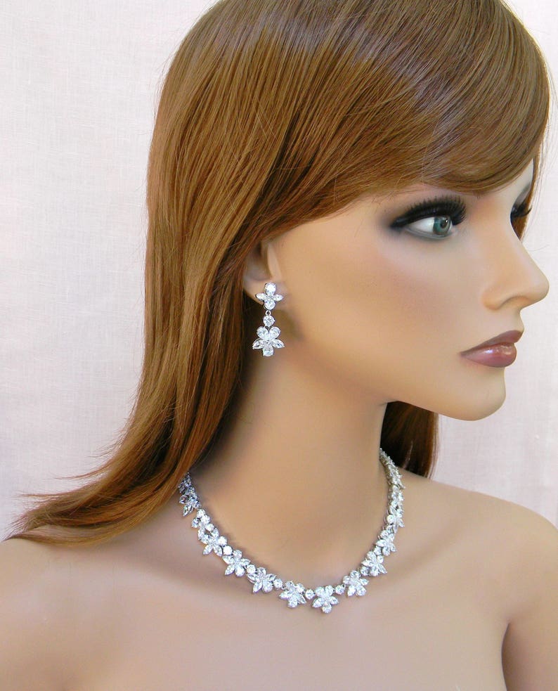 May include: A silver necklace and earrings set with clear crystal flower accents. The necklace is a delicate chain with a row of flower-shaped crystals. The earrings are drop earrings with three flower-shaped crystals.