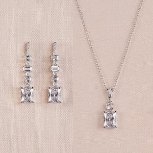 May include: A silver necklace with a square-shaped pendant set with clear cubic zirconia stones. The necklace is paired with matching earrings with a similar design.