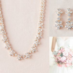 May include: A gold necklace and earrings set with clear crystal flower accents. The necklace is a delicate chain with a floral design. The earrings are long and dangle with a floral design.