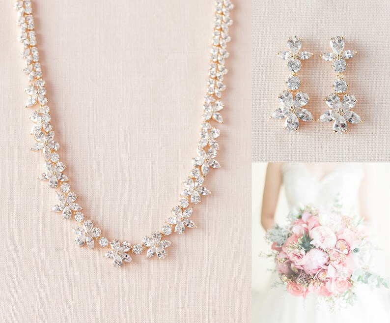 May include: A gold necklace and earrings set with clear crystal flower accents. The necklace is a delicate chain with a floral design. The earrings are long and dangle with a floral design.