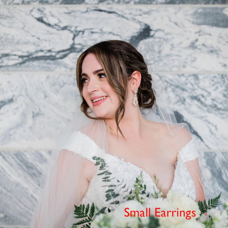 May include: A bride wearing a white off-the-shoulder wedding dress with a veil and small earrings. She is holding a bouquet of white flowers and greenery. The text "Small Earrings" is visible in the image.