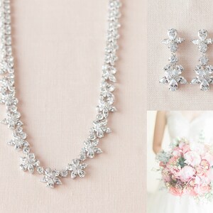 May include: A bridal jewelry set with a silver necklace and earrings. The necklace is made of small, clear stones arranged in a floral pattern. The earrings are also made of clear stones and have a dangling design.
