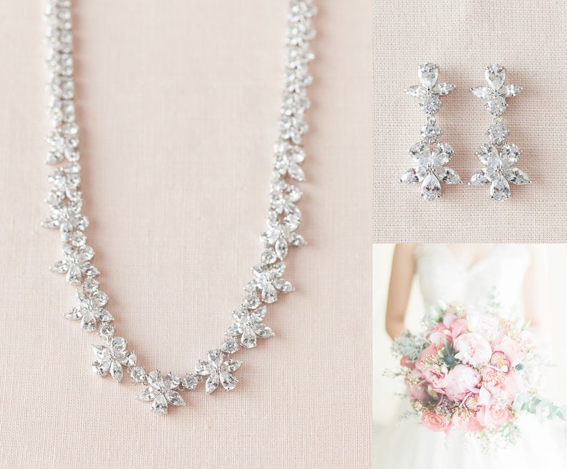 May include: A bridal jewelry set with a silver necklace and earrings. The necklace is made of small, clear stones arranged in a floral pattern. The earrings are also made of clear stones and have a dangling design.