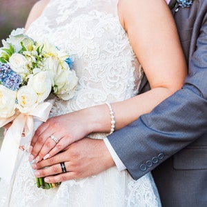 May include: A bride and groom holding hands, the bride is holding a bouquet of white roses and blue flowers, the groom is wearing a gray suit.