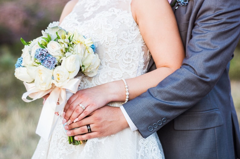 May include: A bride and groom holding hands, the bride is holding a bouquet of white roses and blue flowers, the groom is wearing a gray suit.