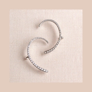 May include: Two silver-toned ear cuffs, each curved in a C-shape, are adorned with small, sparkling crystals. The ear cuffs are displayed on a light beige textured surface, creating a simple yet elegant presentation. These ear cuffs are a stylish jewelry accessory.