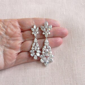 May include: A pair of long, silver dangle earrings with clear crystal and pearl accents. The earrings are teardrop shaped and feature a cluster of crystals at the bottom.