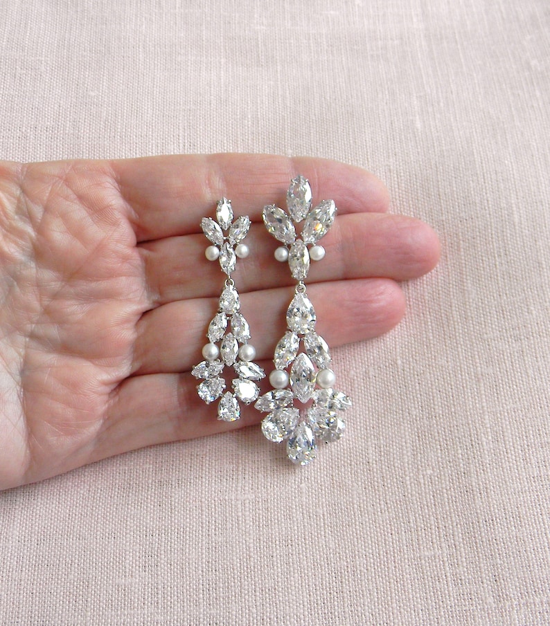 May include: A pair of long, silver dangle earrings with clear crystal and pearl accents. The earrings are teardrop shaped and feature a cluster of crystals at the bottom.