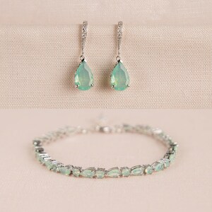 Op de afbeelding: Een set zilveren oorbellen en een armband met druppelvormige groene edelstenen. De oorbellen hebben een delicaat ontwerp en de armband is gemaakt van een dunne ketting.