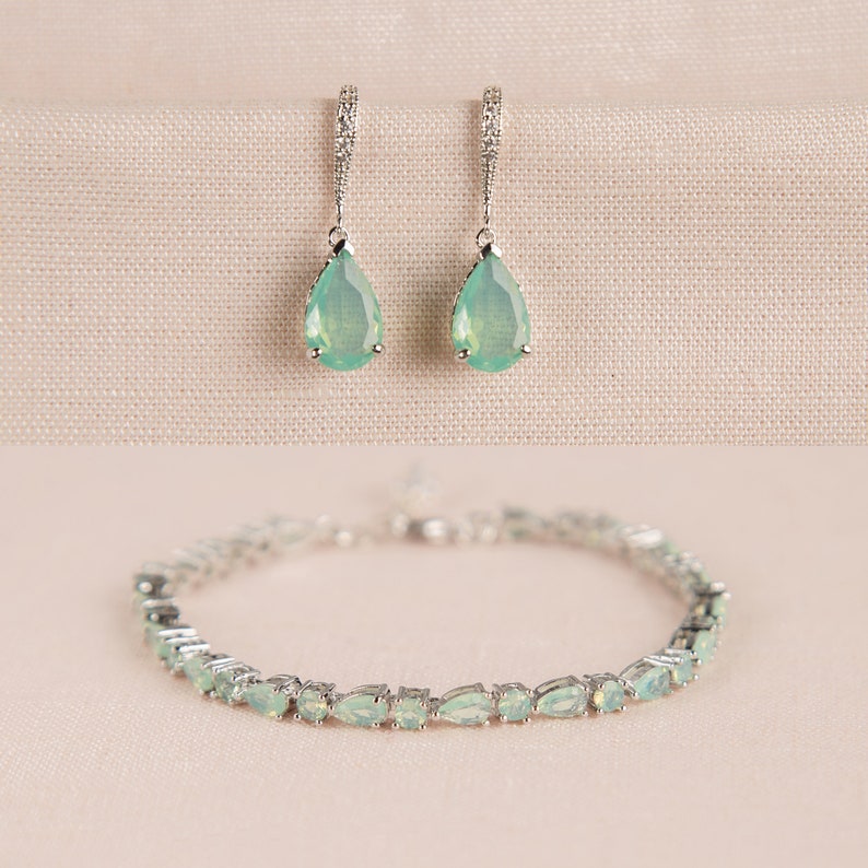 Op de afbeelding: Een set zilveren oorbellen en een armband met druppelvormige groene edelstenen. De oorbellen hebben een delicaat ontwerp en de armband is gemaakt van een dunne ketting.
