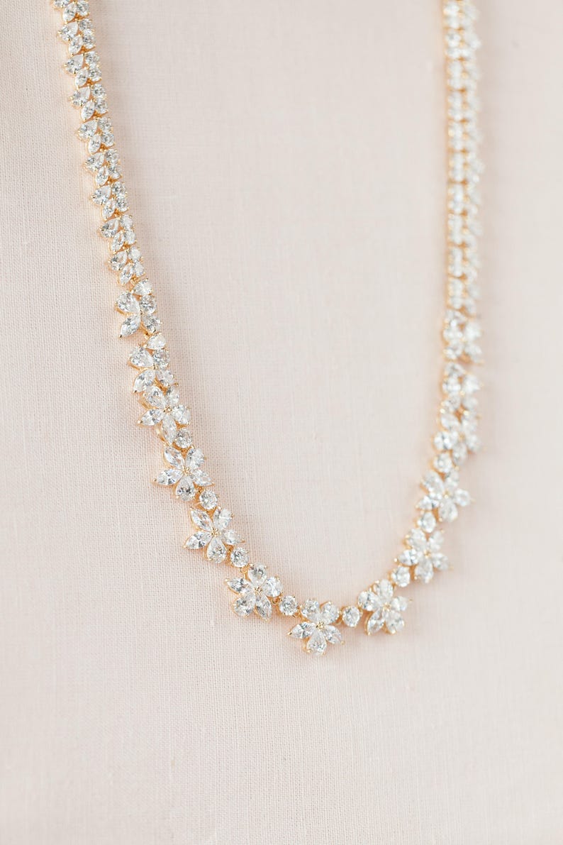 May include: A gold necklace with a delicate design featuring clusters of clear cubic zirconia stones arranged in a floral pattern.