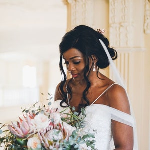 May include: A bride in a white strapless gown with a veil holds a bouquet of pink and green flowers. The bouquet includes proteas, roses, and eucalyptus.
