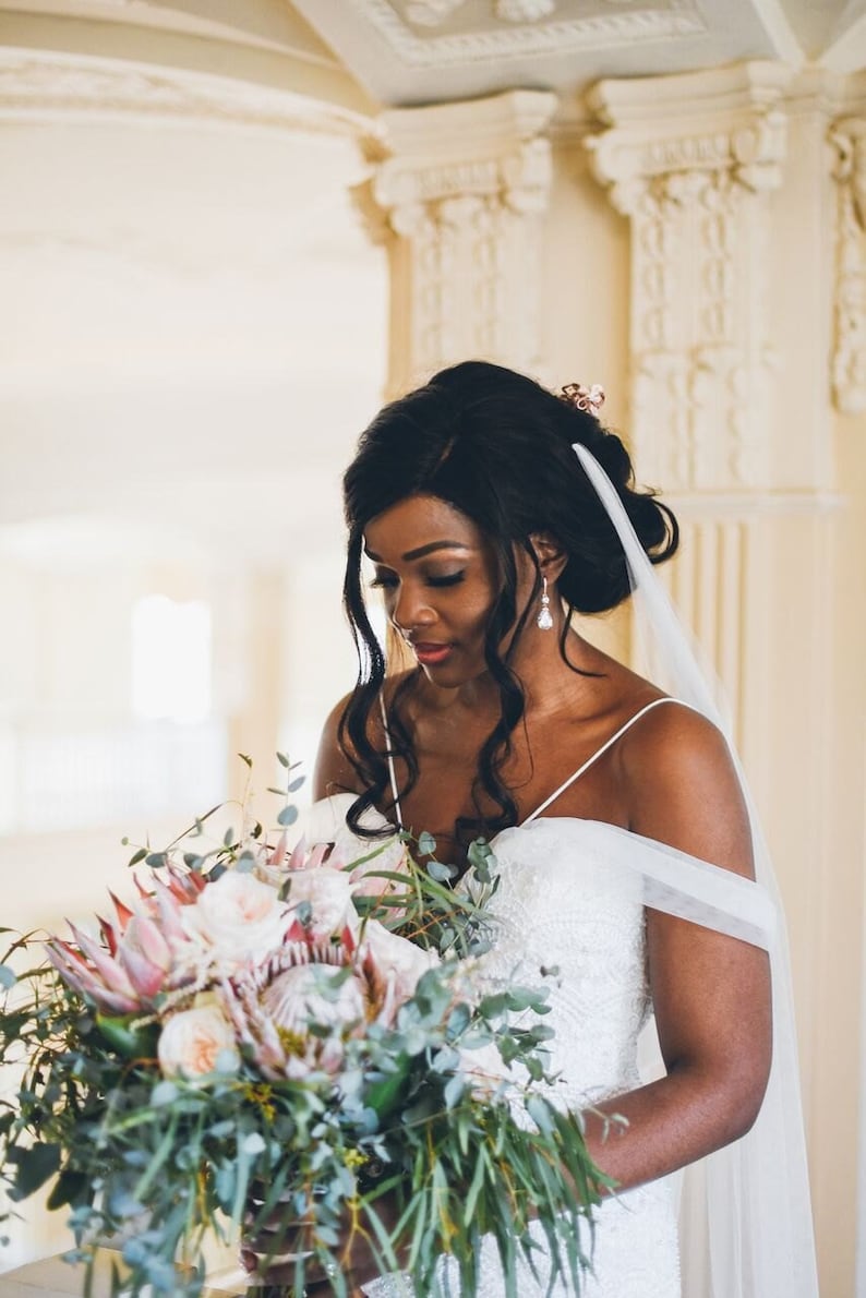 May include: A bride in a white strapless gown with a veil holds a bouquet of pink and green flowers. The bouquet includes proteas, roses, and eucalyptus.