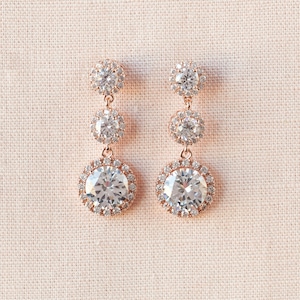 May include: A pair of rose gold dangle earrings with three clear crystal stones. The largest stone is surrounded by a halo of smaller stones.