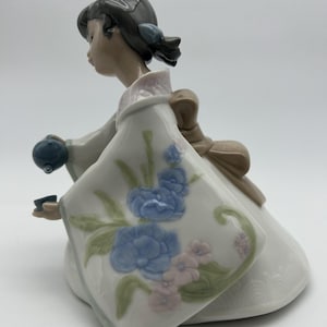 Può includere: Statuina in porcellana di una ragazza in kimono bianco, inginocchiata e con una piccola teiera e tazza blu. Il kimono è decorato con disegni floreali blu e rosa. La ragazza ha i capelli scuri e indossa un fiocco.