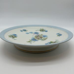 Pode incluir: Um suporte para bolos de cerâmica branca com borda azul clara e design floral. O suporte apresenta uma base elevada com um detalhe dourado. O padrão floral inclui flores azuis, verdes e roxas. Uma peça decorativa.