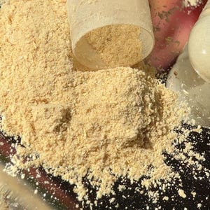 Puede incluir: Primer plano de un polvo de color beige claro que se derrama de un recipiente de pl&aacute;stico transparente. El polvo est&aacute; finamente molido, posiblemente un suplemento o ingrediente. El fondo est&aacute; borroso, con toques de rosa y blanco.