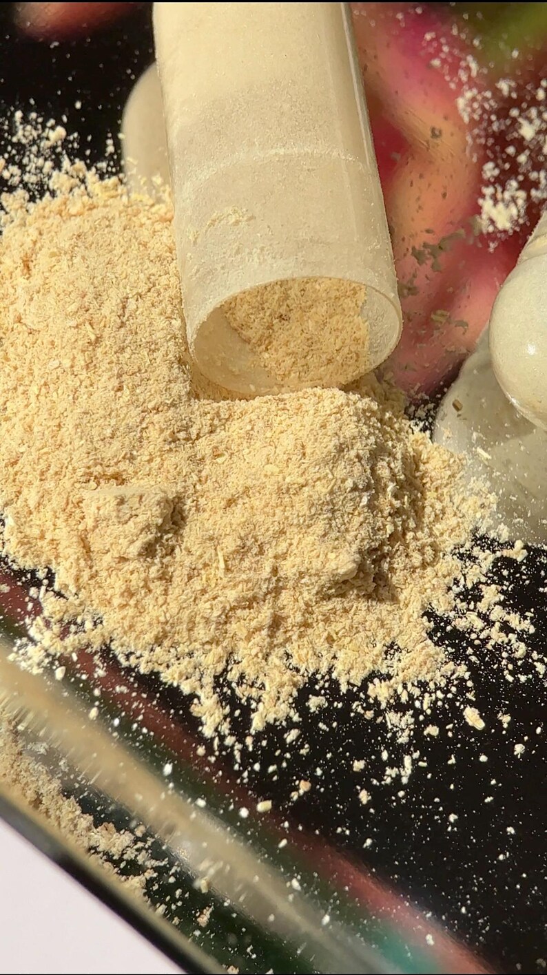 Puede incluir: Primer plano de un polvo de color beige claro que se derrama de un recipiente de pl&aacute;stico transparente. El polvo est&aacute; finamente molido, posiblemente un suplemento o ingrediente. El fondo est&aacute; borroso, con toques de rosa y blanco.