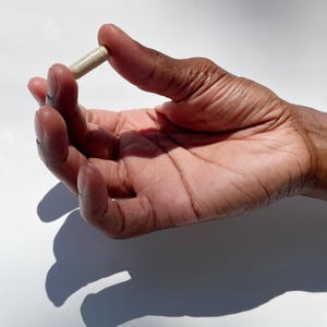 Puede incluir: Una c&aacute;psula beige sostenida entre los dedos de una mano. La mano es de piel oscura con venas y arrugas visibles, sobre un fondo blanco brillante. La c&aacute;psula es cil&iacute;ndrica y parece ser un suplemento o medicamento.