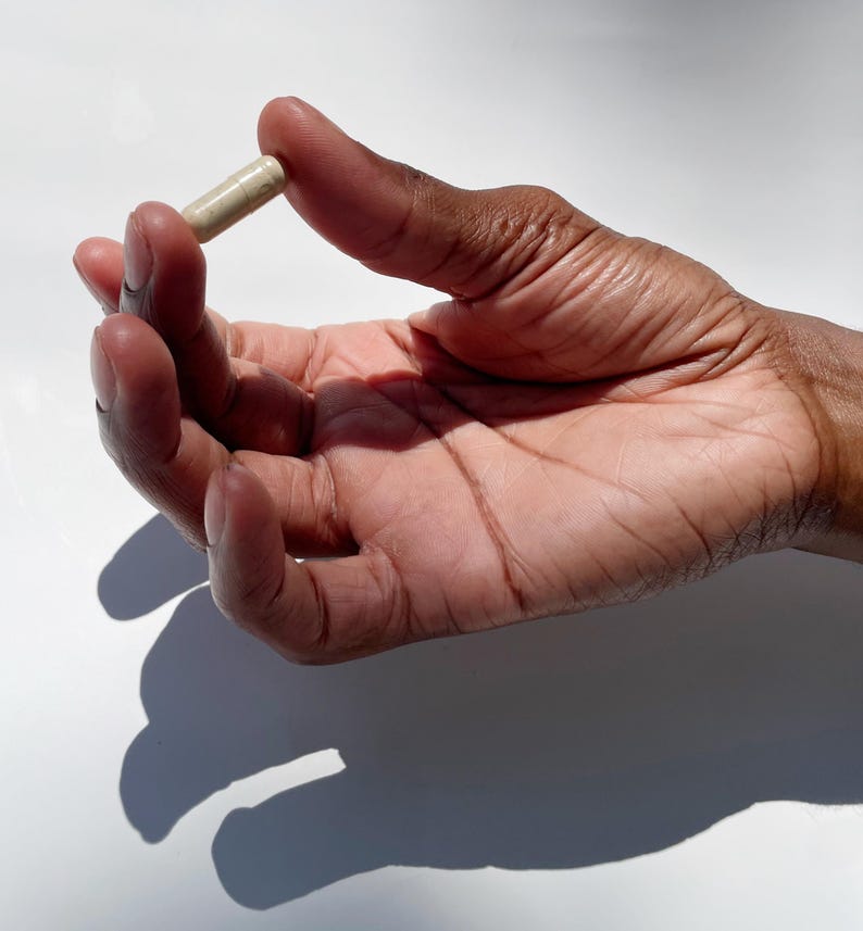 Puede incluir: Una c&aacute;psula beige sostenida entre los dedos de una mano. La mano es de piel oscura con venas y arrugas visibles, sobre un fondo blanco brillante. La c&aacute;psula es cil&iacute;ndrica y parece ser un suplemento o medicamento.