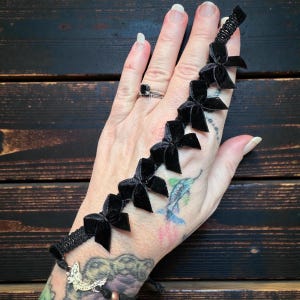 Puede incluir: Pulsera de lazo de terciopelo negro con seis lazos pequeños. La pulsera se muestra en una mano tatuada con un anillo plateado. Los lazos están espaciados uniformemente a lo largo del cordón negro.