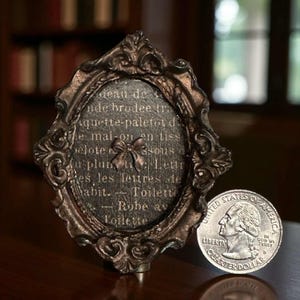 Op de afbeelding: Een antiek ogend, ovaalvormig frame met sierlijke details en een bronzen afwerking. Het frame bevat een gedrukte tekst met een strik illustratie. Een Amerikaanse kwart dollar munt is naast het frame geplaatst. De tekst is in het Frans.