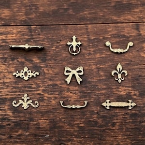 May include: A collection of nine small, antique brass-coloured metal decorative ornaments featuring various shapes including a cross with a ring, a bow, a fleur-de-lis, and several ornate horizontal drawer pull-style hardware pieces.