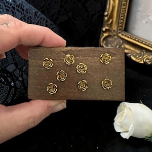 May include: A rectangular dark brown wooden block featuring seven small, metallic gold-colored rose-shaped embellishments arranged across its surface. The rose accents are detailed with layered petals and a polished finish, set against the wood grain.