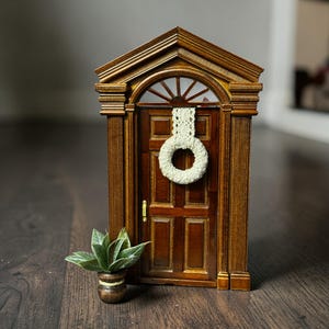 May include: Miniature wooden door with a decorative wreath and a small potted plant. The door is a rich brown color with detailed trim and a small brass handle. The wreath is a light cream color, adding a touch of softness to the scene.