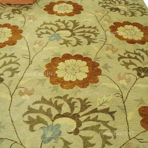Peut inclure: Un grand tapis avec un motif floral. Le tapis a un fond vert clair avec de grandes fleurs stylisées dans des tons de marron, d'orange et de crème. Les fleurs sont reliées par des tiges et des feuilles marron. Le tapis est en matière tissée.