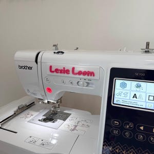 May include: A white Brother SE700 embroidery and sewing machine with the name "Lexie Loom" in pink lettering. The machine has a digital display and various buttons. The machine is on a wooden surface.